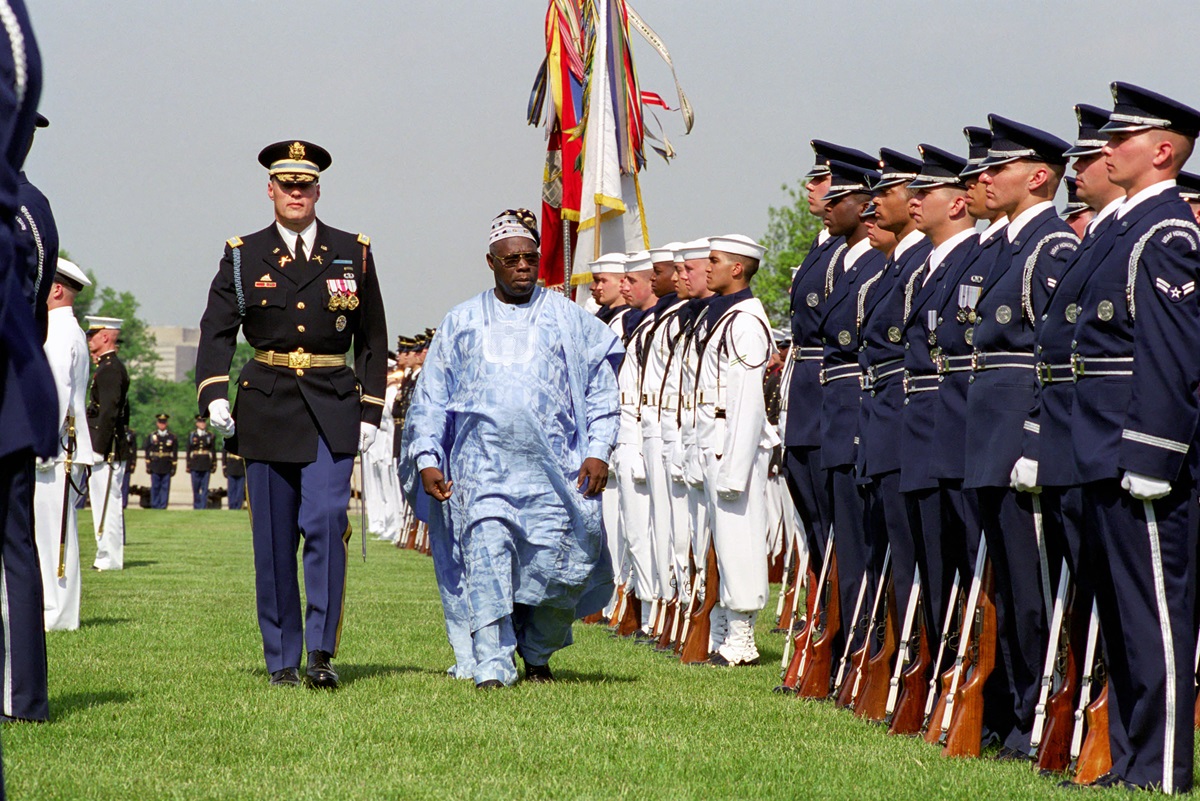 Obasanjo - THEWILL NEWS MEDIA Military parade: a man in blue traditional attire walks between rows of sailors in dress uniforms during a ceremony on a grass field with flags in the background.