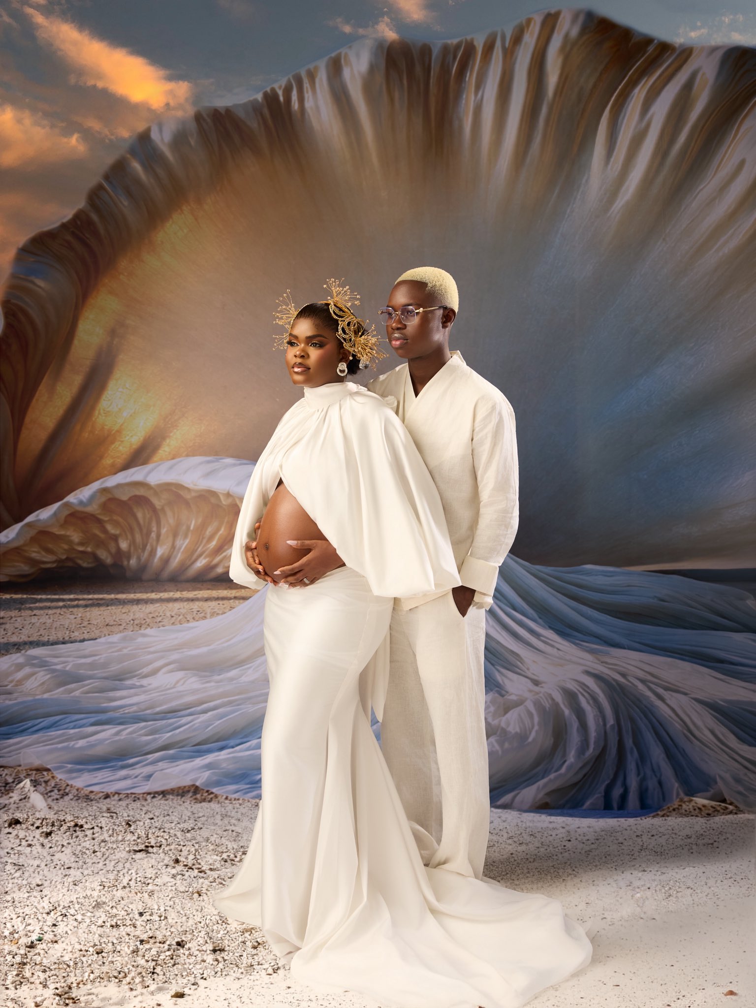 Peller and wife Jarvis - THEWILL NEWS MEDIA Pregnant woman in a white gown cradles her belly while a partner in white stands behind her on a sandy beach with a flowing, sculptural backdrop behind them.