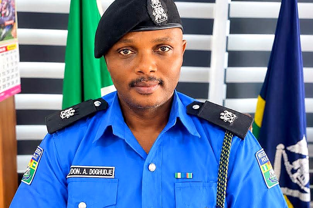 SP Johnson A Doghudje - THEWILL NEWS MEDIA Police officer in a bright blue uniform with a black beret, name tag reads 'Don. A. Doghudje', posed indoors with flags in the background.