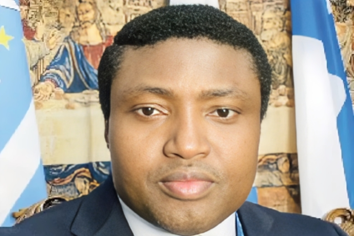Simon Ekpa - THEWILL NEWS MEDIA Head-and-shoulders portrait of a man in a dark suit with a neutral expression, blue and white flags in the background.