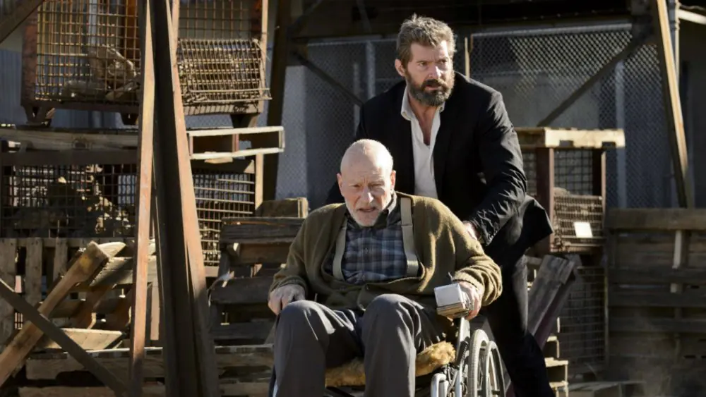 Sir Patrick Stewart - THEWILL NEWS MEDIA An elderly man in a wheelchair is being pushed by a bearded man in a dark suit through a scrapyard with wooden crates and metal cages nearby.