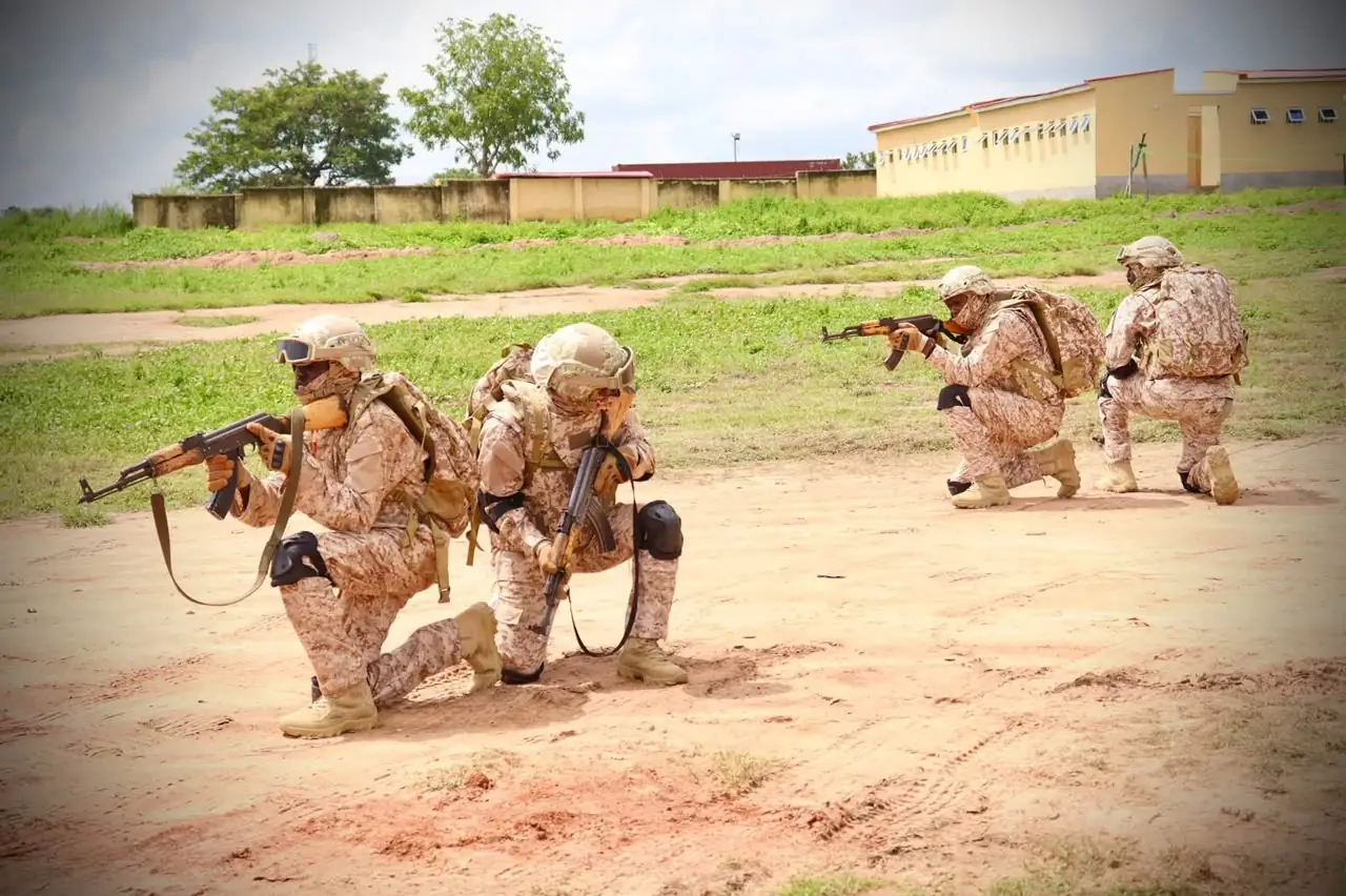 Four soldiers in desert camouflage kneel and aim rifles during training on a dusty field with buildings in the background.