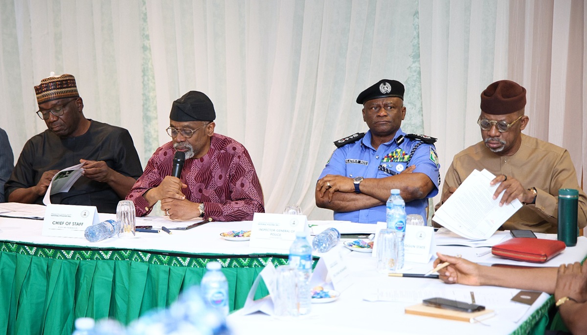 State policing - THEWILL NEWS MEDIA Panel of officials seated at a long table during a formal meeting; a man in traditional attire speaks into a microphone while others, including a police officer, listen and review documents on the table