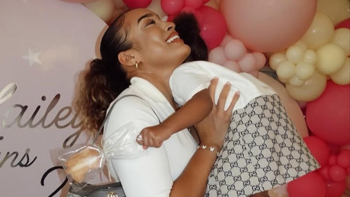 Stephanie Ladewig and daughter - THEWILL NEWS MEDIA Pregnant woman in white smiles while hugging a baby, with pastel pink and peach balloons in the background.