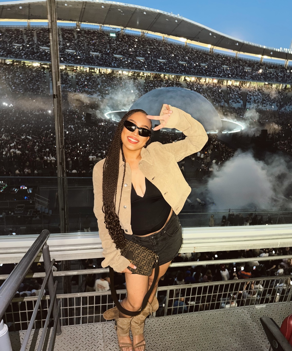 Stephanie Ladewig in stadium - THEWILL NEWS MEDIA Smiling woman with long braids and sunglasses posing on a stadium railing, beige jacket over a black top, with a packed arena and smoky stage behind.