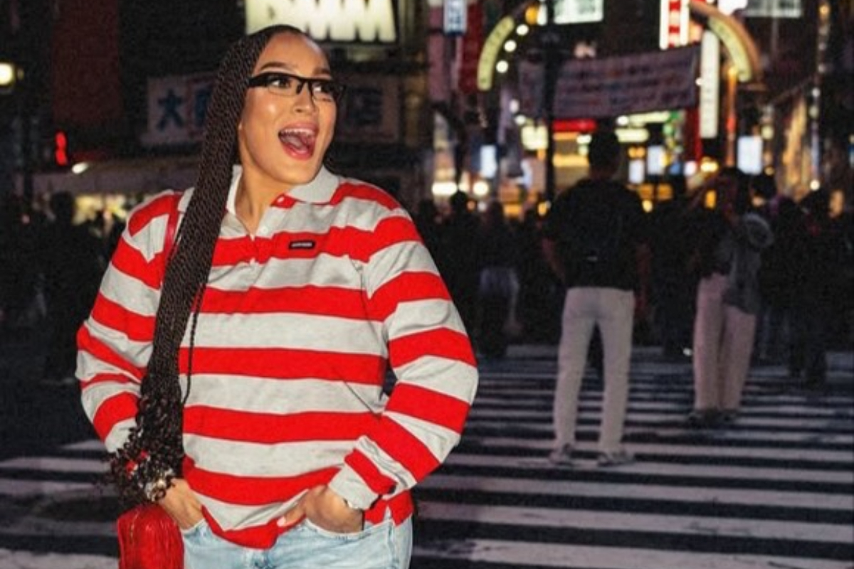 Stephanie Ladewig - THEWILL NEWS MEDIA Woman with braided hair and glasses in a red-and-white striped shirt on a crosswalk at night in a busy city street.