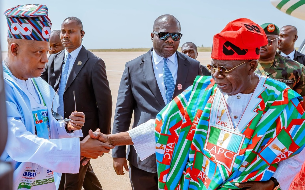 Tinubu and Shettima - THEWILL NEWS MEDIA Two men shake hands on an airstrip; one in light blue traditional attire, the other in a colorful APC outfit with a red cap, surrounded by security.