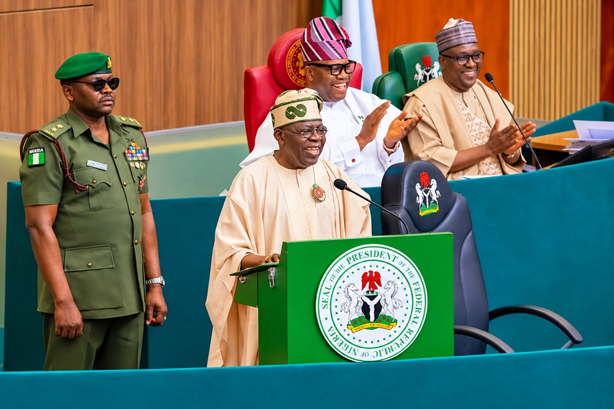 Tinubu with Akpabio and Abass - THEWILL NEWS MEDIA President addressing a formal gathering from a green podium bearing the Nigerian seal, with three officials seated behind.