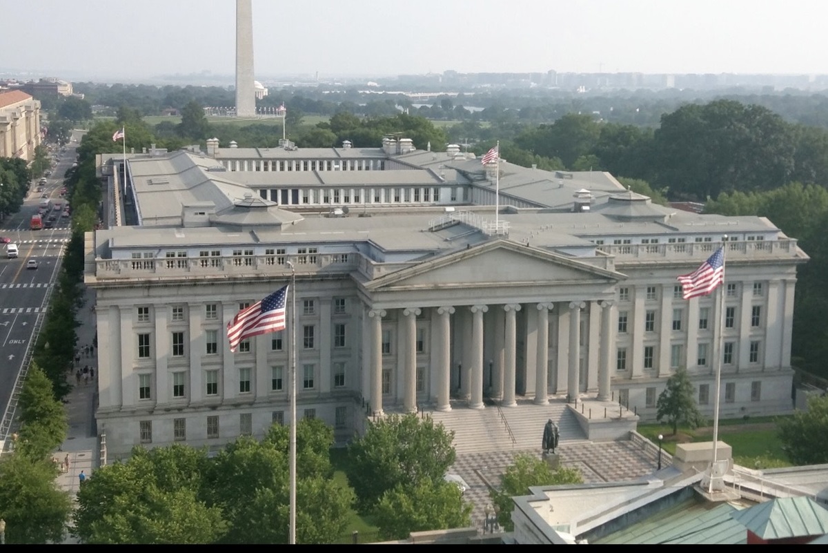US Treasury building in Washington DC - THEWILL NEWS MEDIA A large neoclassical government building with a columned portico, American flags, and a tiled plaza in front, surrounded by trees and streets seen from above.
