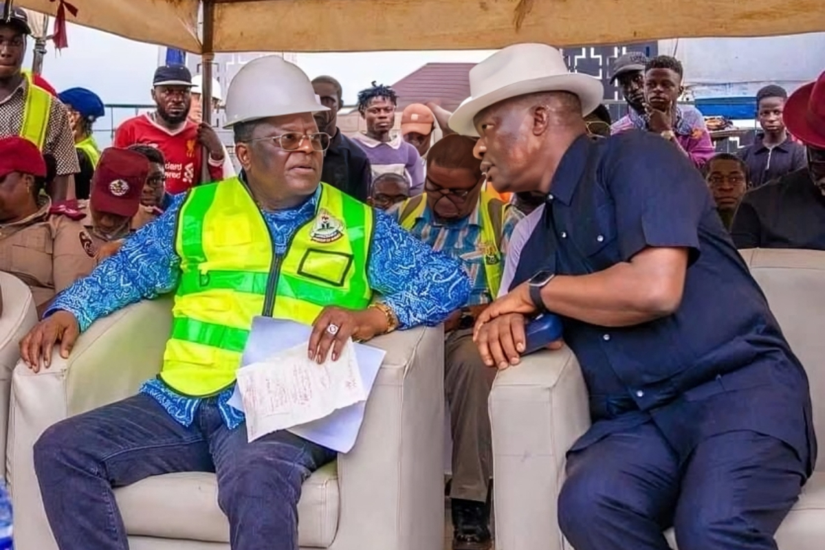 Umahi at First Niger Bridge - THEWILL NEWS MEDIA Two men sit and talk during a construction-site event; one in a neon safety vest and white hard hat holds papers, the other in a dark blue outfit and white hat leans in, surrounded by onlookers.