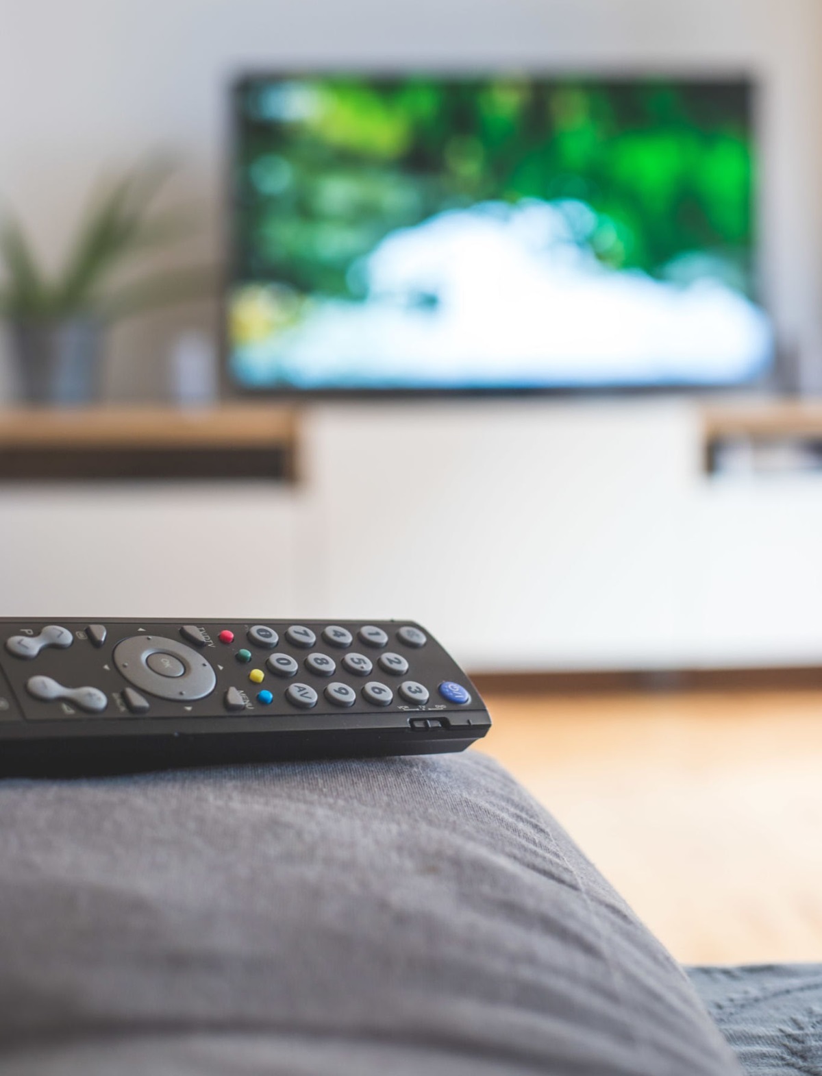 TV remote control in the foreground tv in the blurry background Streaming - THEWILL NEWS MEDIA Black TV remote resting on a gray couch/sofa fabric with a blurred living room and a wall-mounted TV in the background