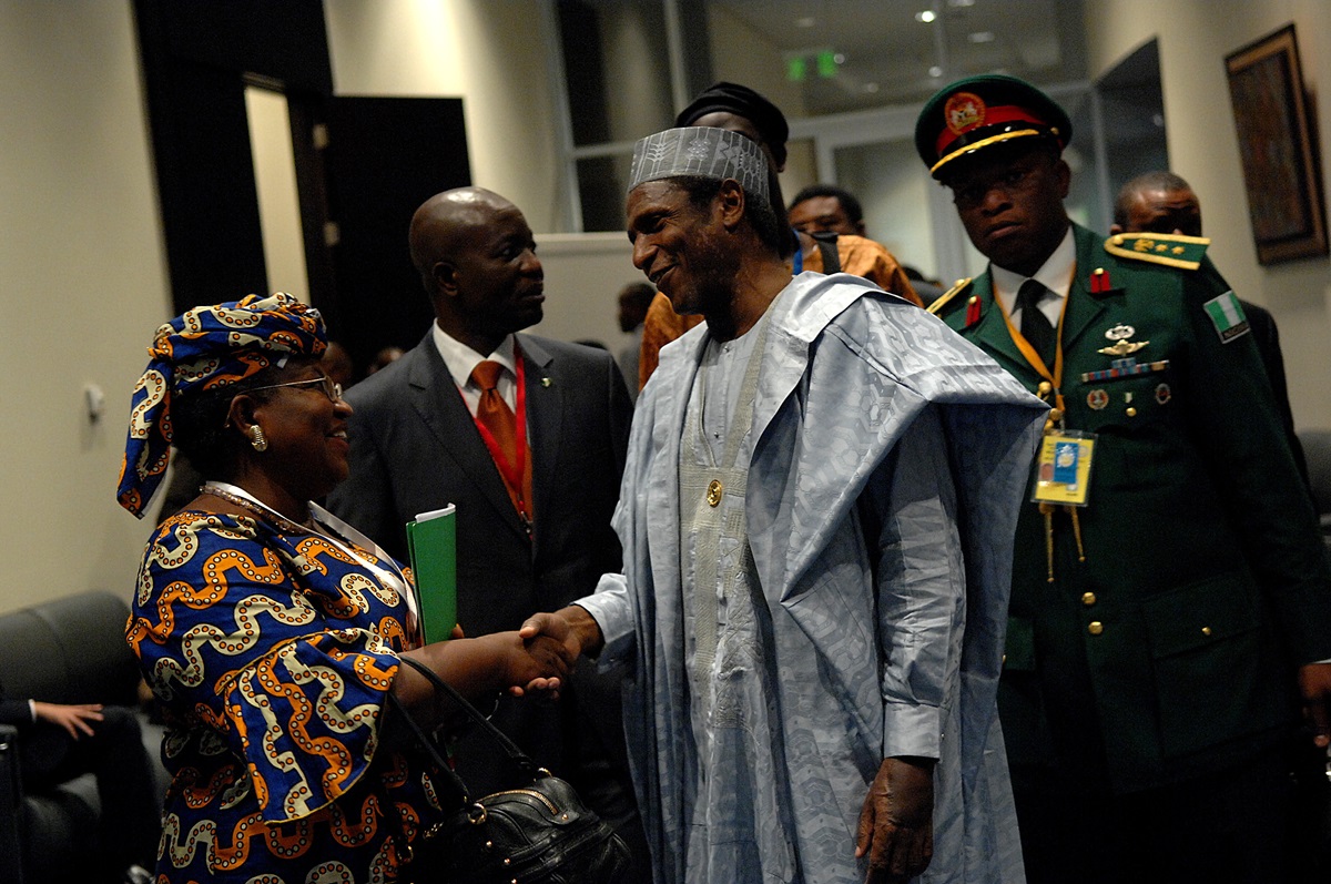 Yaradua - THEWILL NEWS MEDIA President wearing traditional light blue attire shakes hands with a woman in colorful patterned dress in a hallway, others look on.