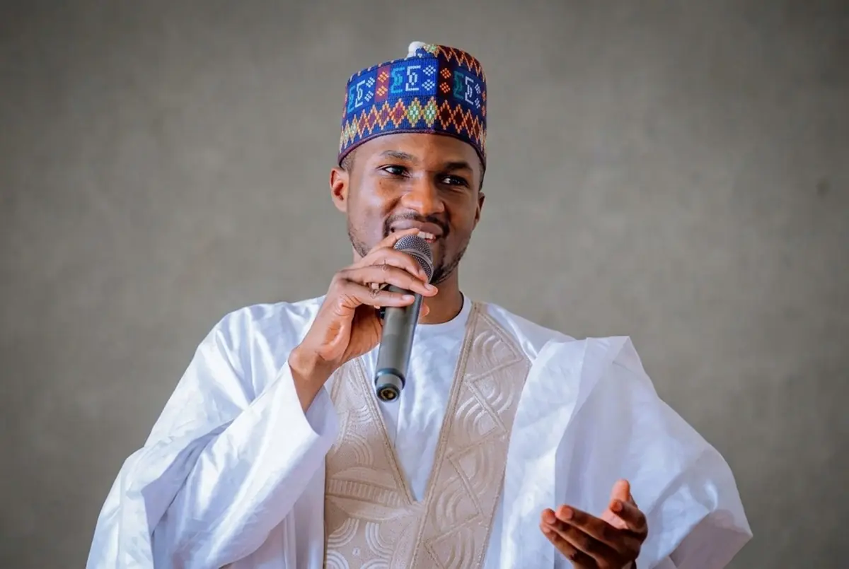 Yusuf Buhari - THEWILL NEWS MEDIA Man in a white traditional outfit and colorful cap speaks into a handheld microphone, likely delivering a speech or performance, against a neutral backdrop.