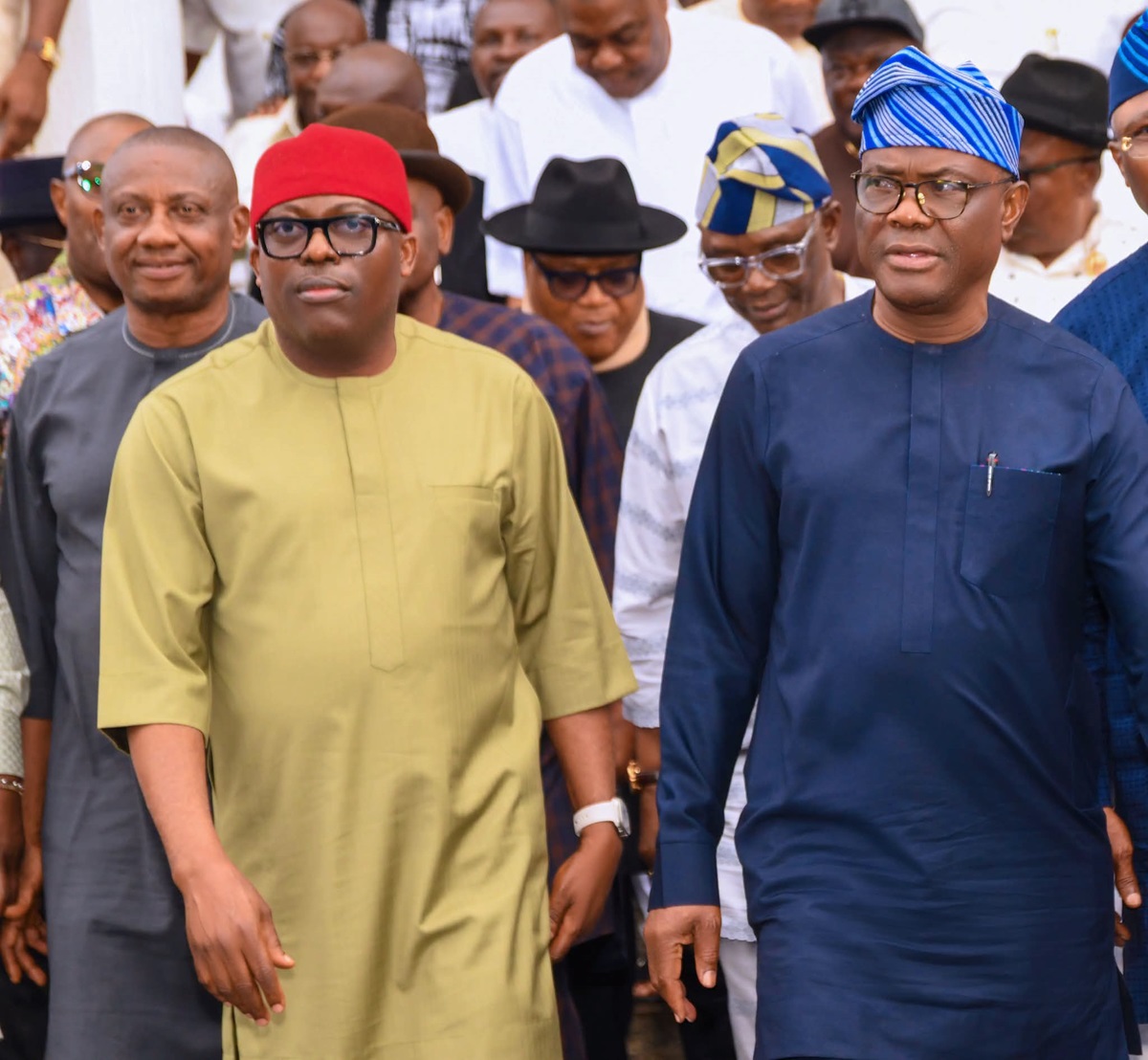 wike and fubara - THEWILL NEWS MEDIA Two men in traditional attire lead a crowd at a formal gathering; one wears an olive outfit with a red cap, the other a navy blue outfit with a blue patterned cap.